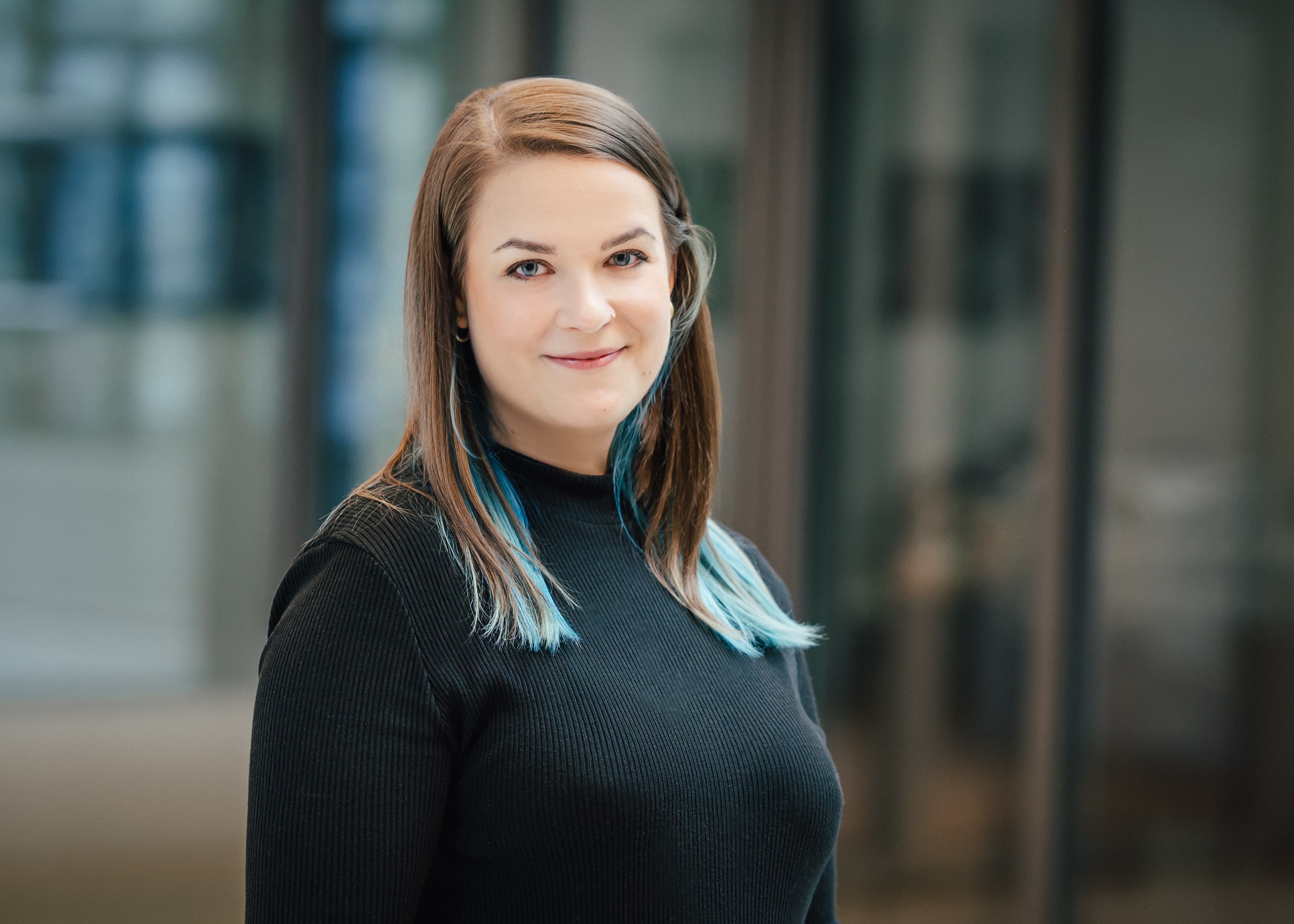 Frau mit langen braunen Haaren und blauen Strähnen, trägt schwarzes Oberteil, Porträtaufnahme vor unscharfem Hintergrund..